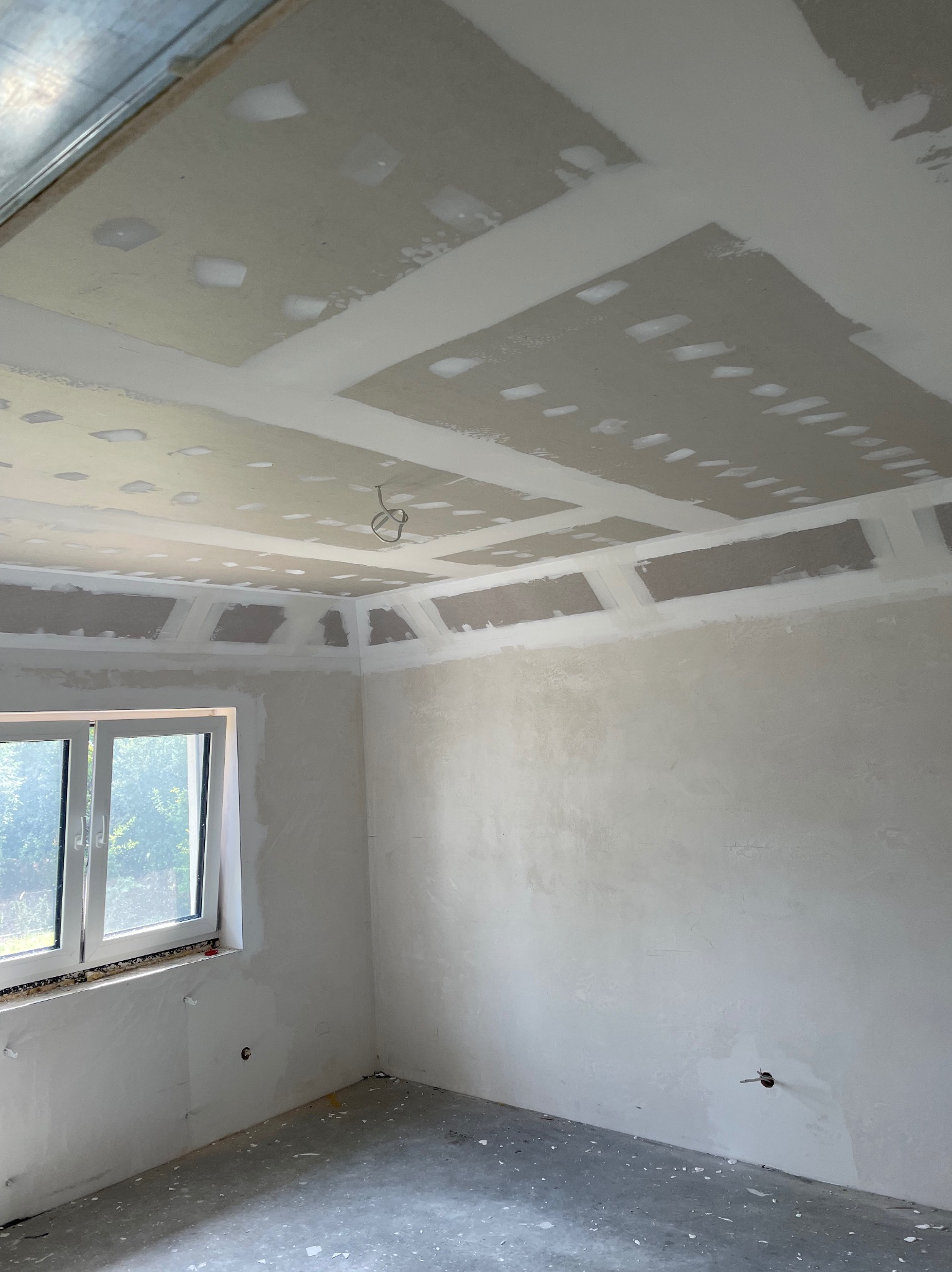 Wnętrze w trakcie prac wykończeniowych, widoczny sufit z płyt kartonowo-gipsowych z widocznymi łączeniami, częściowo pokryty masą szpachlową, ściany przygotowane do dalszych prac, okno z widokiem...