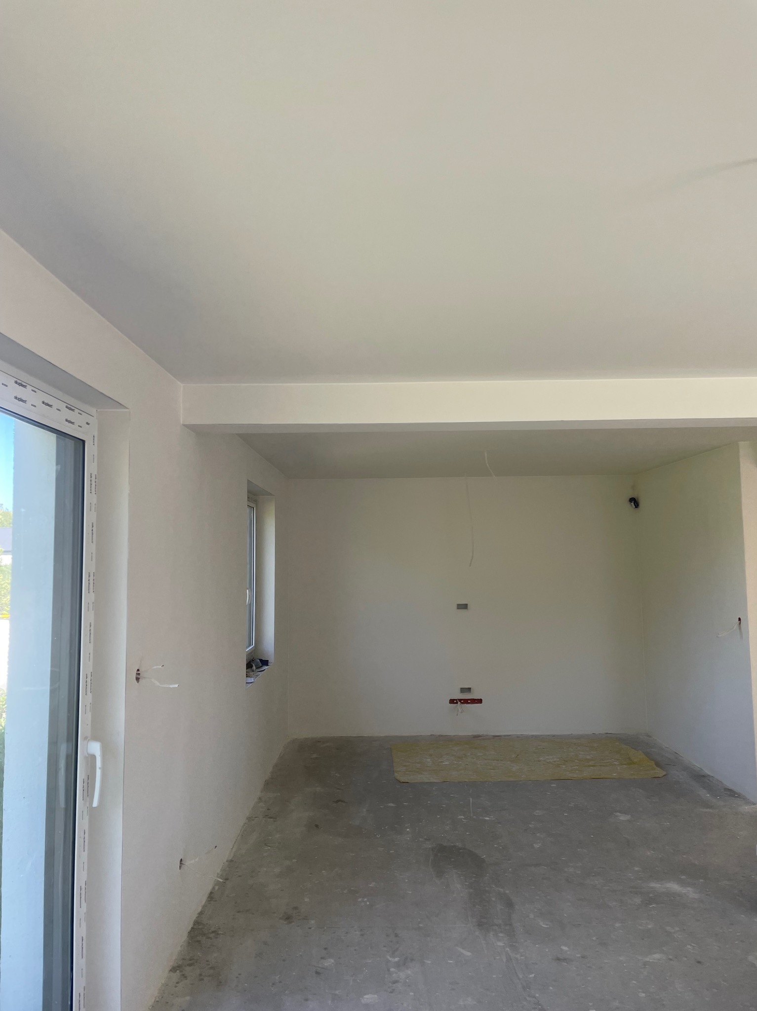 Surowe wnętrze w trakcie prac wykończeniowych, widoczne ściany po nałożeniu gładzi, surowa podłoga, wystające kable elektryczne, okno z widokiem na zieleń.