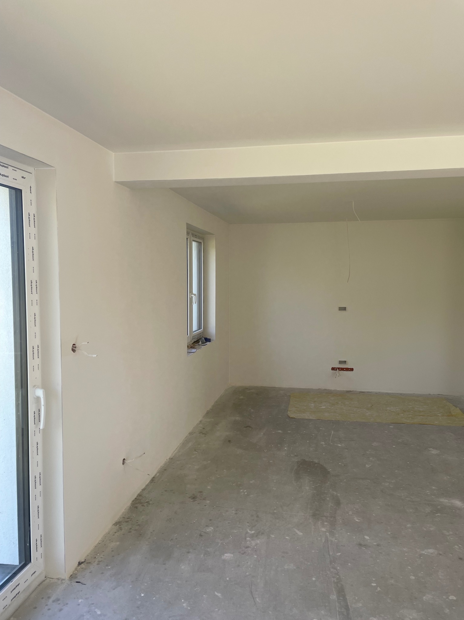 Wnętrze w stanie deweloperskim z oknami PCV, świeżo wygładzonymi ścianami i surowym betonem na podłodze, przygotowane do dalszych prac wykończeniowych.