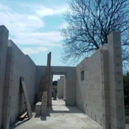 Wznoszenie ścian domu z bloczków betonowych, widoczne otwory okienne i drzwiowe, na zewnątrz widok na drzewo bez liści i błękitne niebo.