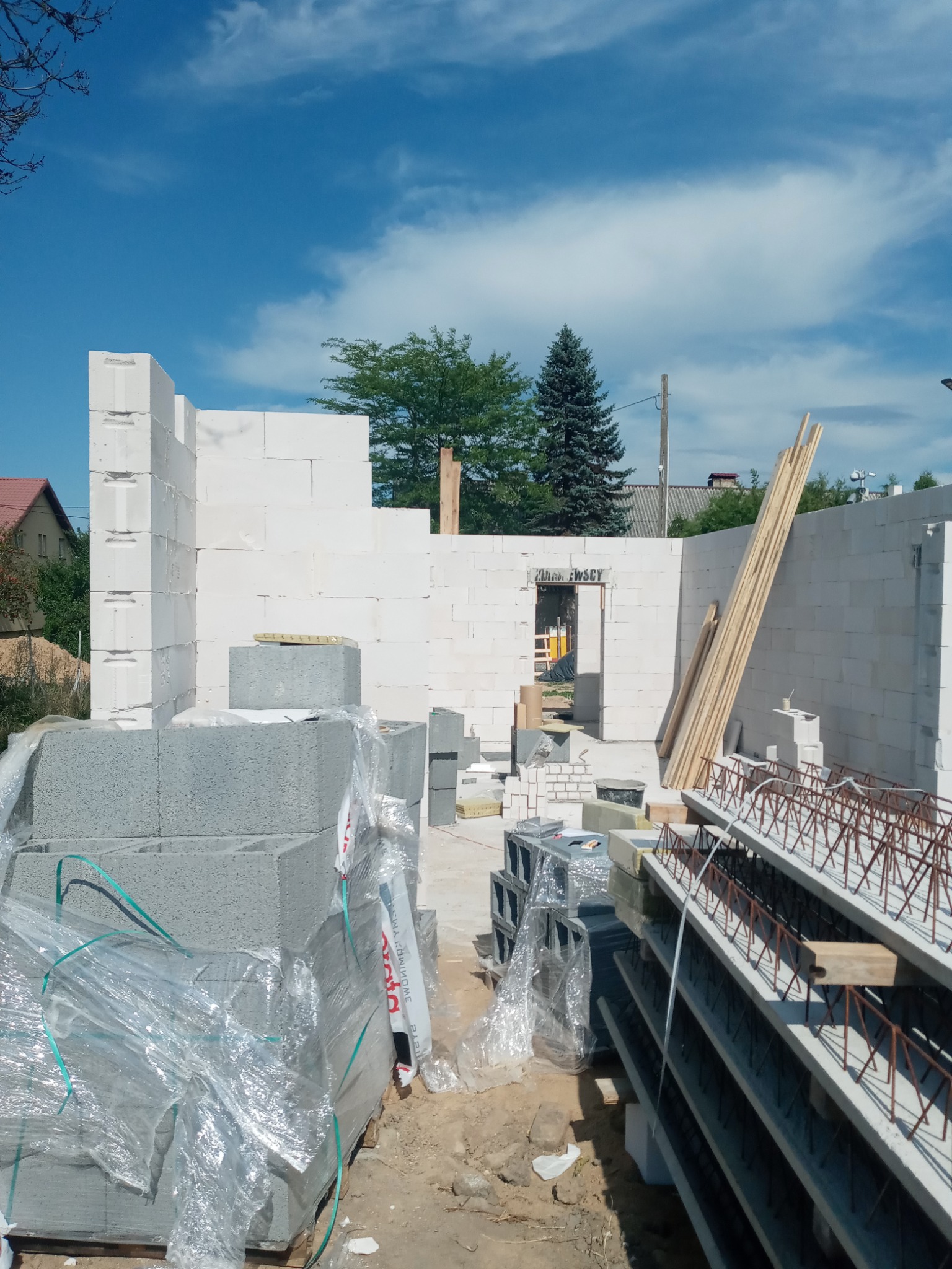 Budowa domu w trakcie: ściany z białych bloczków, stosy bloczków szarych w folii, belki stropowe zbrojone, drewniane deski, błękitne niebo.