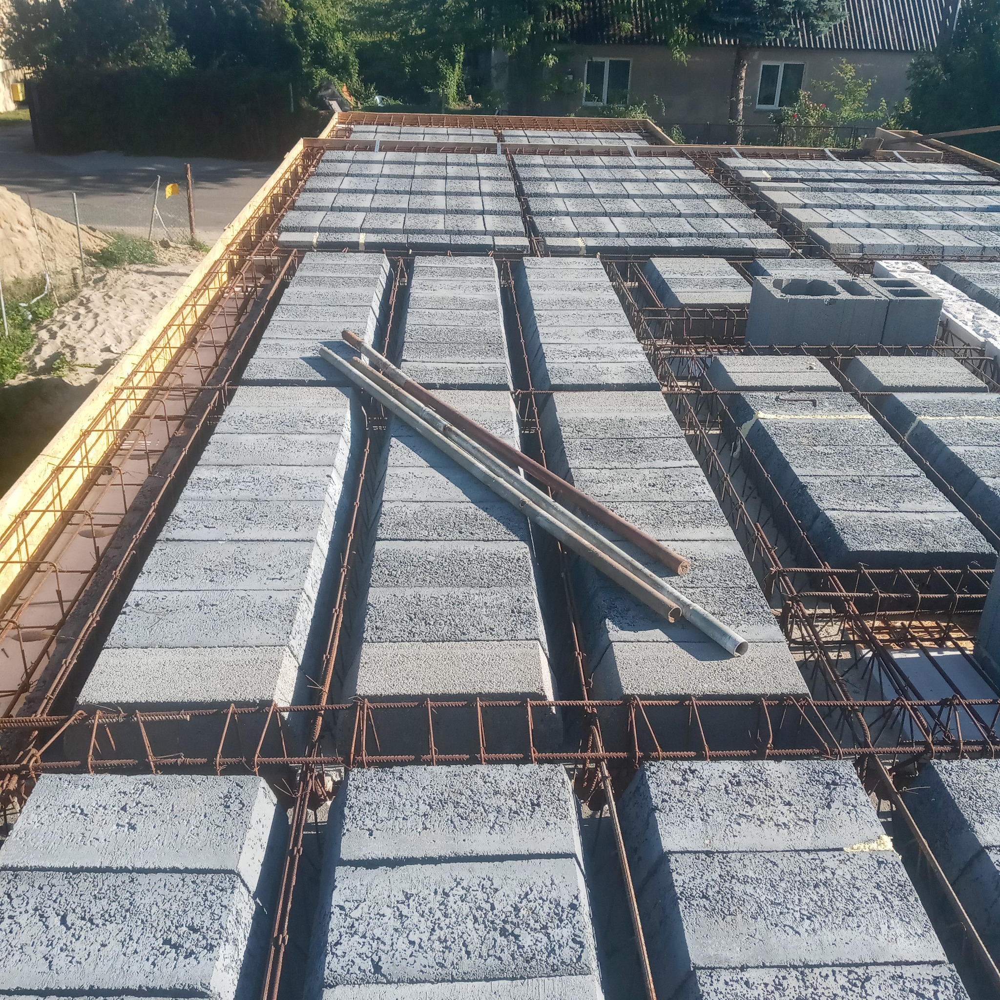 Budowa stropu Teriva: pustaki betonowe ułożone na deskowaniu z widoczną stalową siatką zbrojeniową, trzy rury leżące na pustakach, budynek w tle.