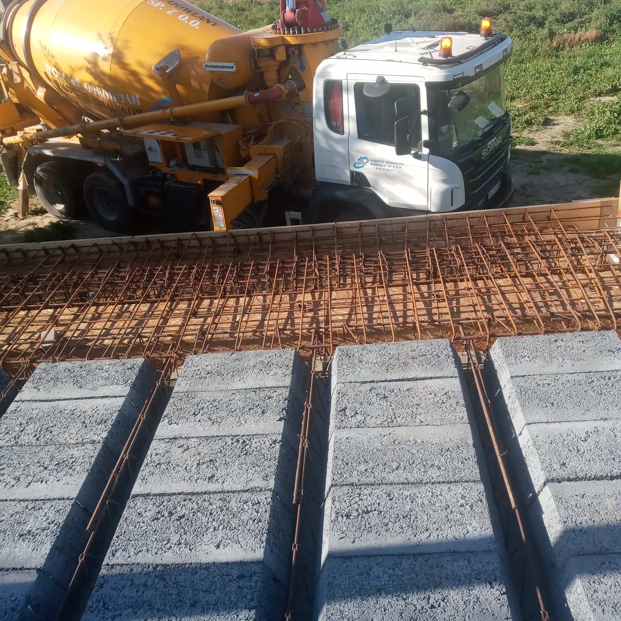 Betoniarka Putzmeister obok zbrojenia stropu z pustaków szalunkowych, widoczne detale zbrojenia i pustaków.