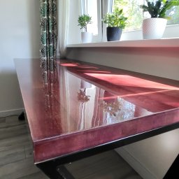 LIONTECH Jakub Kiedrowski - Stół z blatem z żywicy epoksydowej w odcieniach czerwieni, wsparty na czarnej metalowej konstrukcji, w tle okno z roślinami doniczkowymi i wzorzystymi zasłonami.