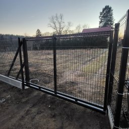 LIONTECH Jakub Kiedrowski - Czarna, przesuwna brama wjazdowa z metalowej siatki panelowej, zamontowana na betonowej podstawie z widoczną szyną prowadzącą, na tle pola i drzew.