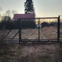 Dwuskrzydłowa brama wjazdowa z czarnej siatki stalowej, widok z zewnątrz posesji, z widocznymi śladami opon na błotnistym podłożu i domem z czerwonym dachem w tle o zmierzchu.