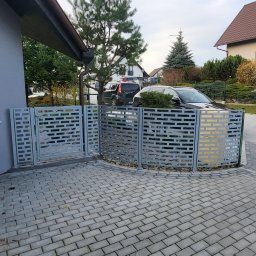 LIONTECH Jakub Kiedrowski - Szare, łukowe ogrodzenie z ażurowym wzorem, dopasowane do kostki brukowej. Brama wjazdowa z identycznym wzorem. W tle domy i samochody.