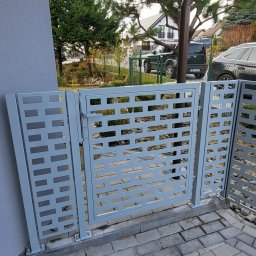 LIONTECH Jakub Kiedrowski - Szara, nowoczesna brama wjazdowa z ażurowym wypełnieniem z prostokątnych otworów, zamontowana na posesji z kostką brukową. Widok z bliska, w tle zieleń i budynki.