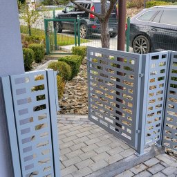 LIONTECH Jakub Kiedrowski - Nowoczesna, szara brama wjazdowa z ażurowym wzorem, zamontowana na posesji z kostką brukową i zielenią. Widoczne detale mocowania i wykończenia.