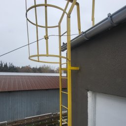 LIONTECH Jakub Kiedrowski - Żółta drabina ewakuacyjna z koszem ochronnym, zamontowana na ścianie budynku z szarą elewacją. Widok z dołu, na tle pochmurnego nieba i zabudowań przemysłowych.