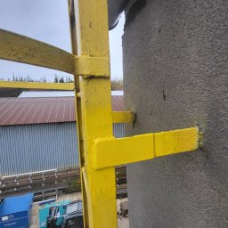 LIONTECH Jakub Kiedrowski - Żółta, metalowa drabina przymocowana do szarej ściany budynku. W tle widoczny wózek widłowy i magazyn z blachy falistej. Ujęcie z dołu, podkreślające wysokość konstrukcji.