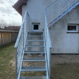 LIONTECH Jakub Kiedrowski - Zewnętrzne schody metalowe z balustradą, ocynkowane ogniowo, prowadzące do wejścia na piętro budynku. Stopnie z kraty WEMA, filary i słupki balustrady z profili stalowych.