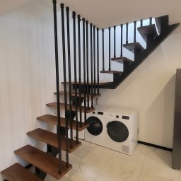LIONTECH Jakub Kiedrowski - Stalowe schody z drewnianymi stopniami i czarną balustradą w nowoczesnym wnętrzu. Widoczne pralki w tle. Ujęcie z dołu, podkreślające konstrukcję.