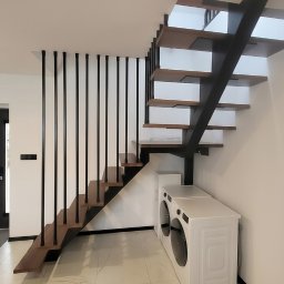 LIONTECH Jakub Kiedrowski - Nowoczesne schody metalowe z drewnianymi stopniami i czarną balustradą w minimalistycznym wnętrzu. Widoczne pralki pod schodami. Ujęcie z dołu, podkreślające konstrukcję.