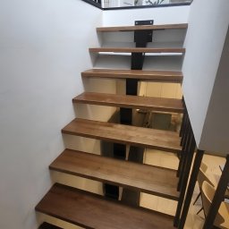 LIONTECH Jakub Kiedrowski - Drewniane schody z metalową konstrukcją i czarną balustradą w nowoczesnym wnętrzu. Stopnie z wyraźnym usłojeniem, centralna belka i minimalistyczny design.
