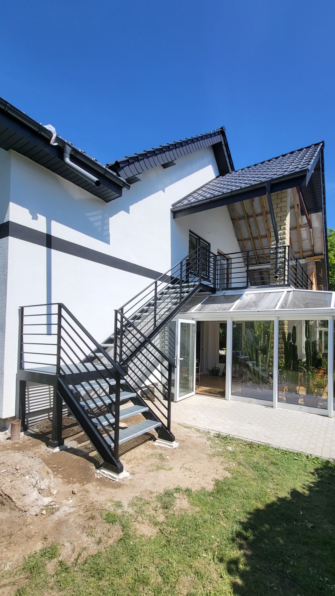 Zewnętrzne schody metalowe z czarną balustradą prowadzą na balkon z przeszkloną zabudową. Widoczne stopnie z kraty WEMA i profilowane słupki. Dom z białą elewacją.