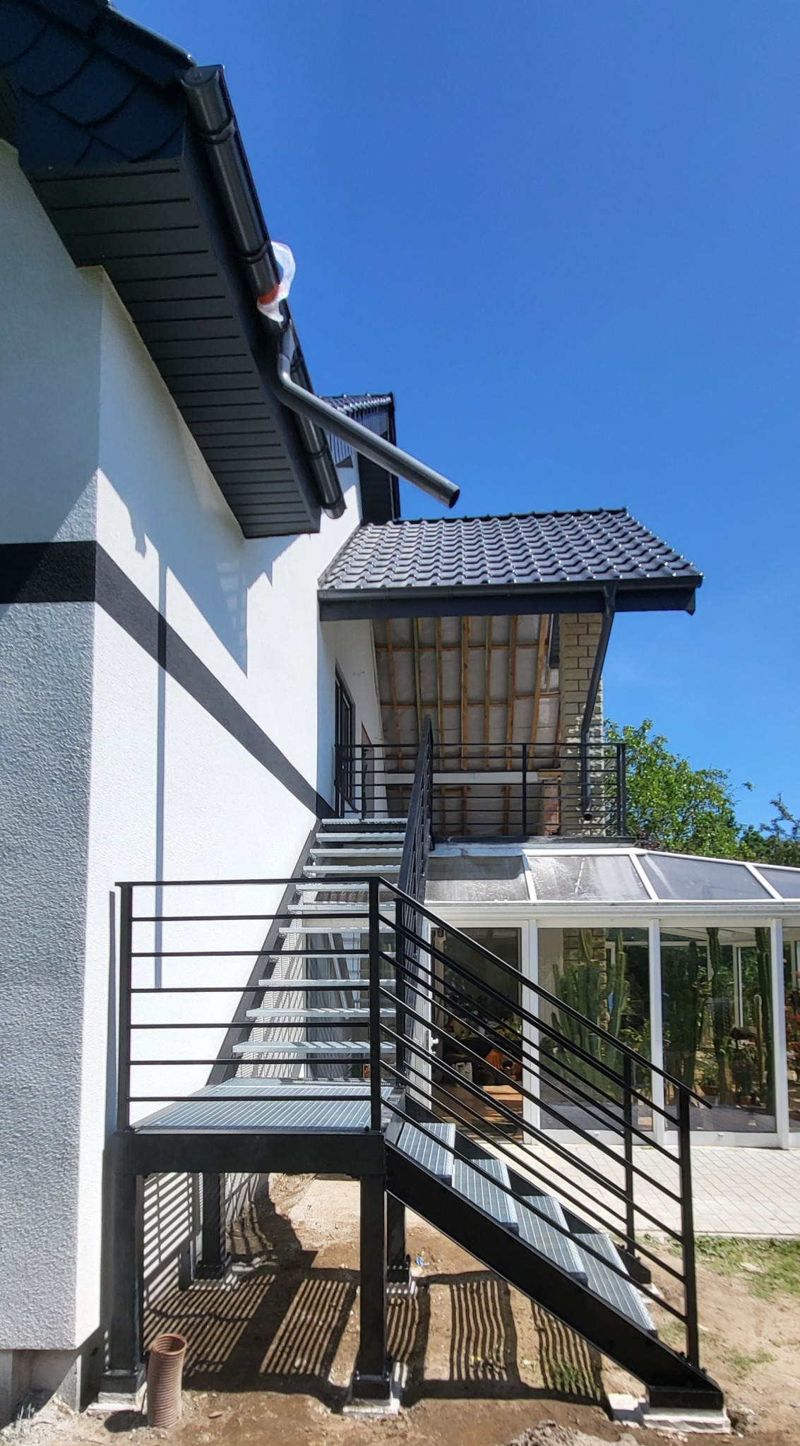 Zewnętrzne schody metalowe z balustradą, prowadzące do przeszklonego ogrodu zimowego. Konstrukcja ocynkowana i pomalowana proszkowo na czarno. Widok z dołu, błękitne niebo.