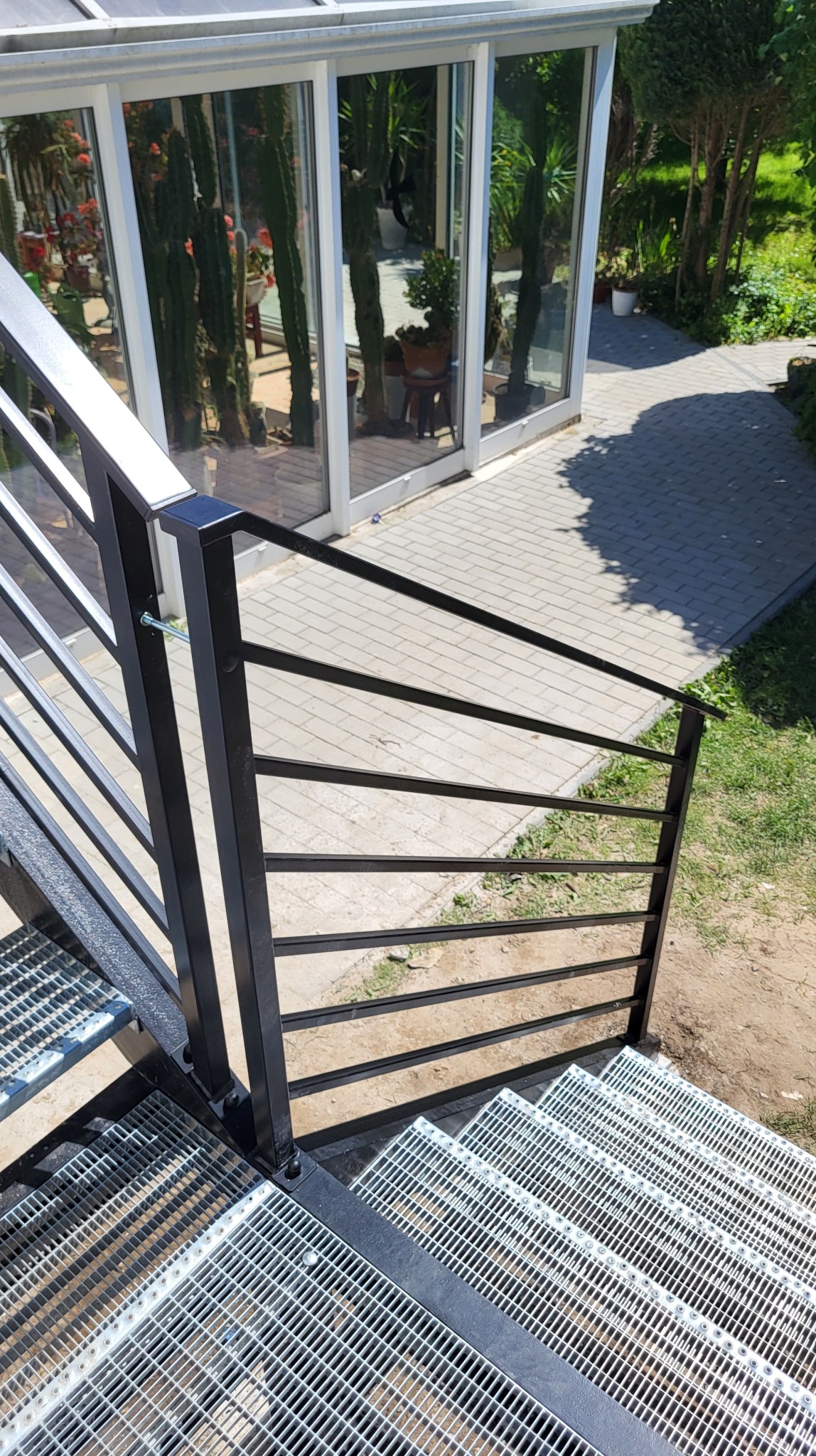Metalowe schody zewnętrzne z czarną, minimalistyczną balustradą z poziomych profili, prowadzące do przeszklonej oranżerii z egzotyczną roślinnością.