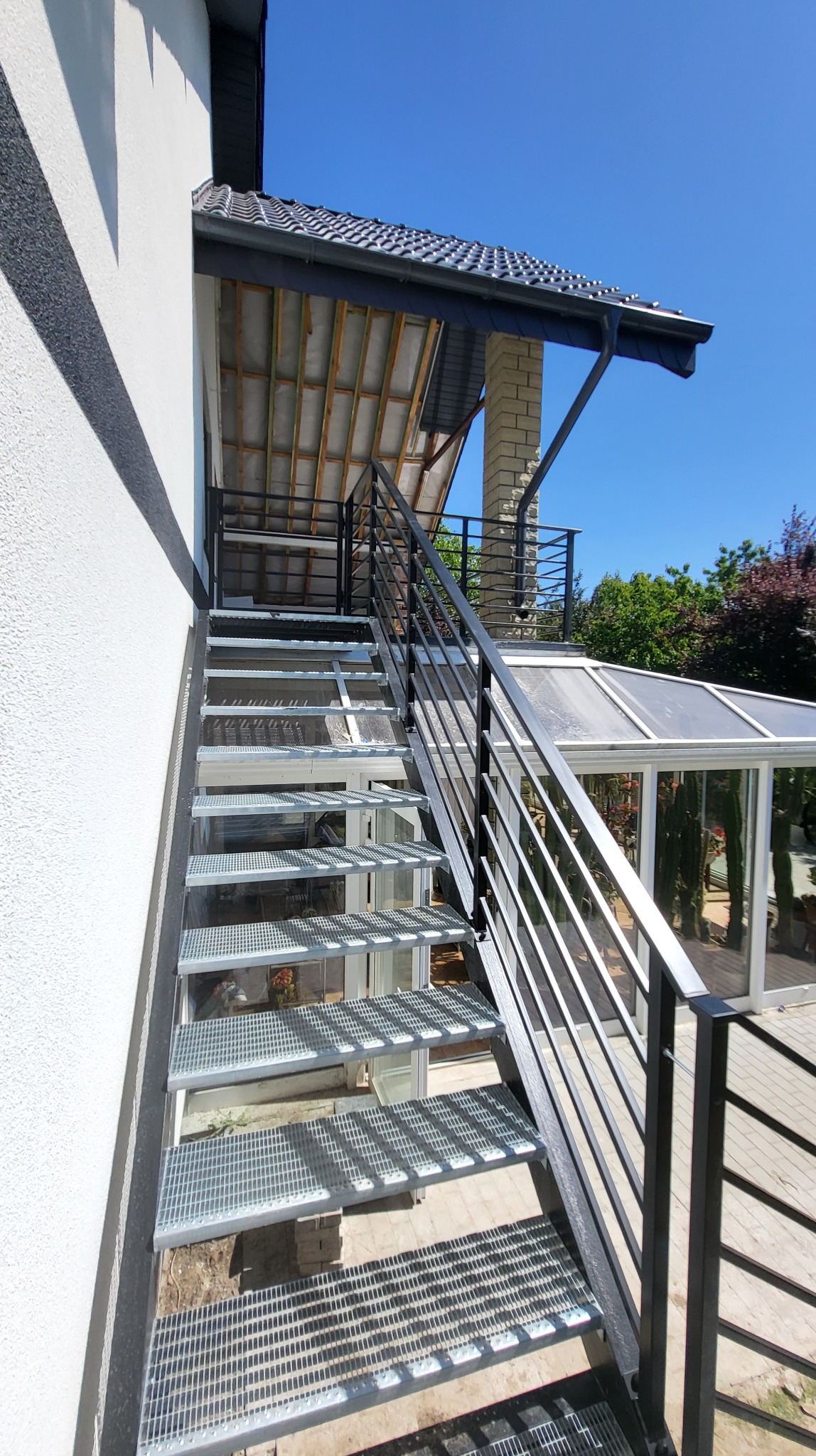 Zewnętrzne schody metalowe z czarną balustradą, ocynkowane i malowane proszkowo, prowadzące do przeszklonej konstrukcji, na tle błękitnego nieba.