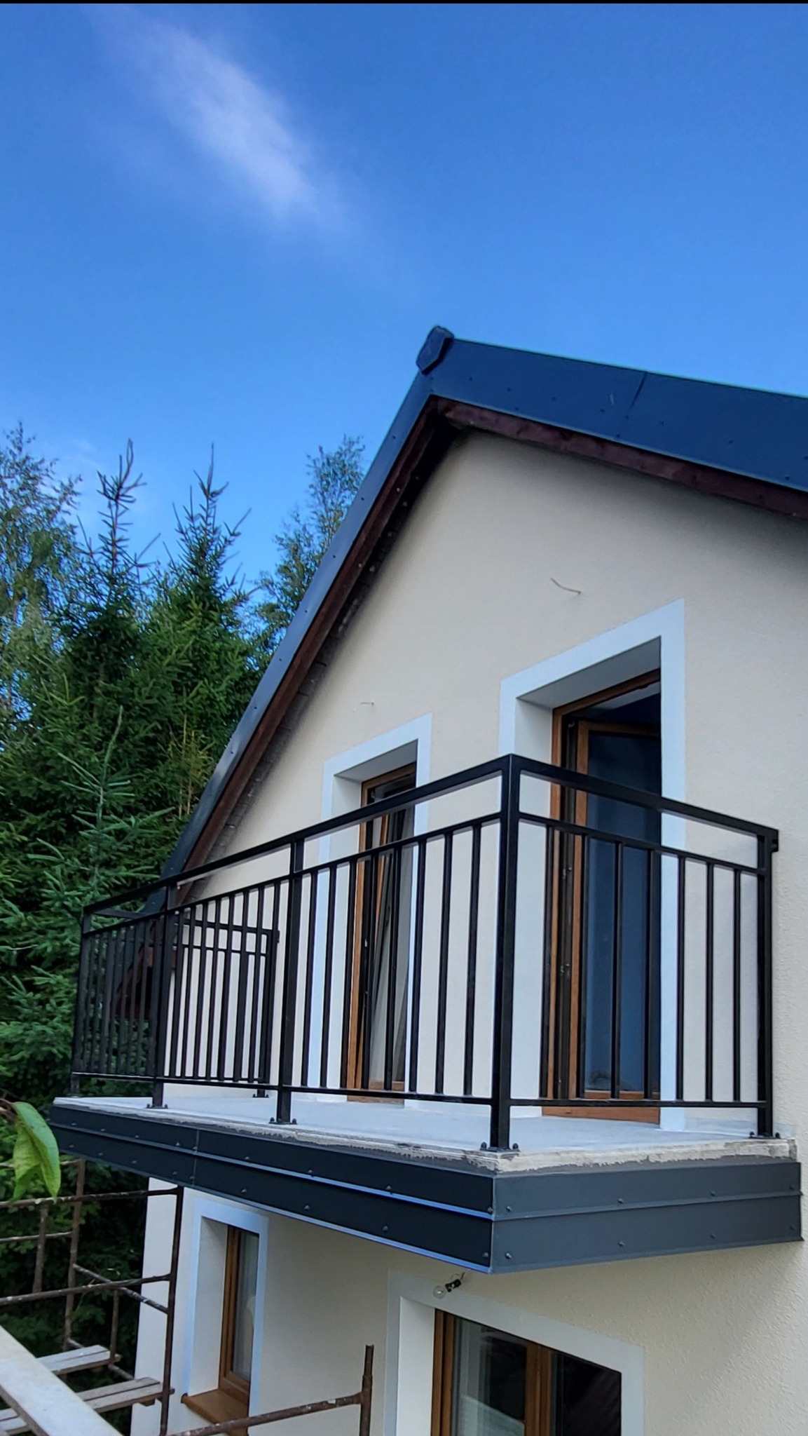 Czarna, metalowa balustrada balkonowa z pionowym wypełnieniem, mocowana do ściany budynku. Widok z dołu, błękitne niebo w tle.