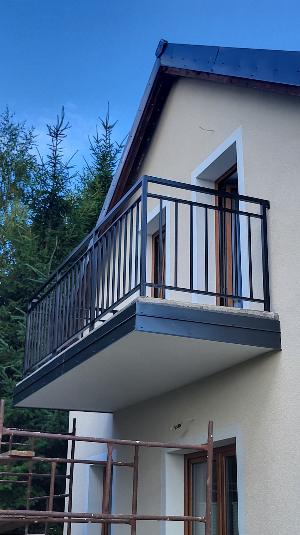 Czarna, metalowa balustrada balkonowa z pionowym wypełnieniem, zamontowana na jasnym budynku z widocznymi fragmentami rusztowania i zieleni w tle.