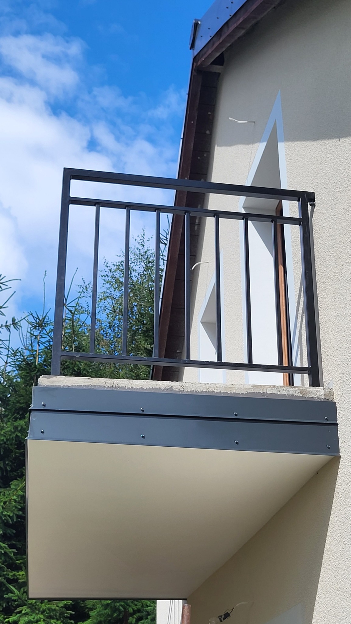 Czarna, metalowa balustrada balkonowa z pionowym wypełnieniem z profili, pomalowana proszkowo, widoczna na tle budynku i błękitnego nieba.