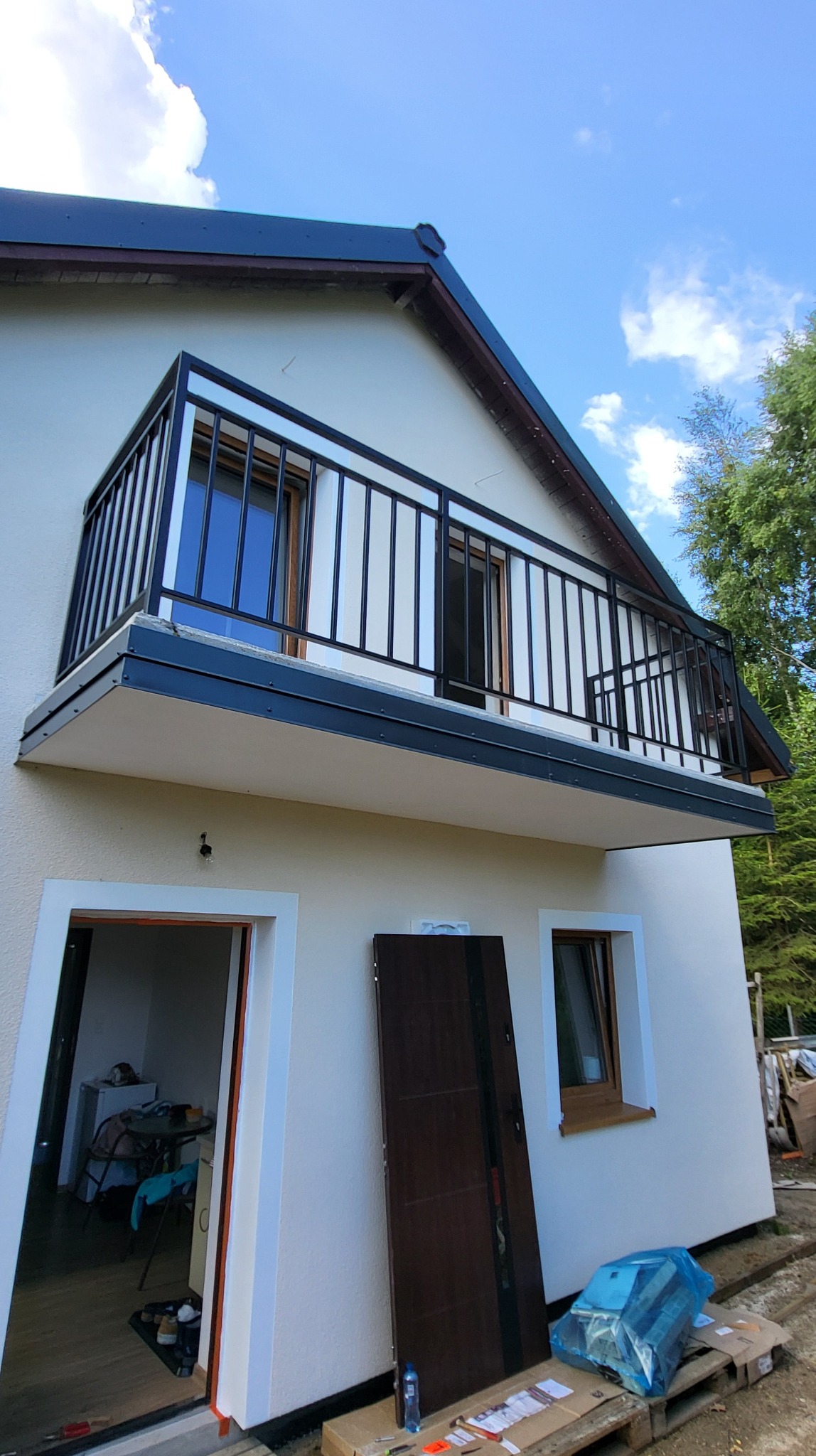 Czarna, metalowa balustrada balkonowa z pionowymi profilami, zamontowana na balkonie domu z białą elewacją, widok z dołu w słoneczny dzień.