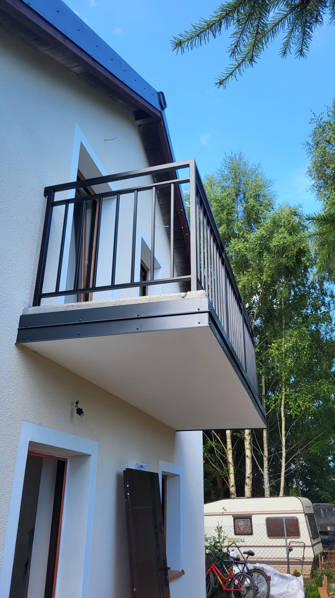 Czarna, metalowa balustrada balkonowa z pionowym wypełnieniem, zamontowana przy budynku z widokiem na zieleń i przyczepę kempingową w tle, wykonana w nowoczesnym stylu.