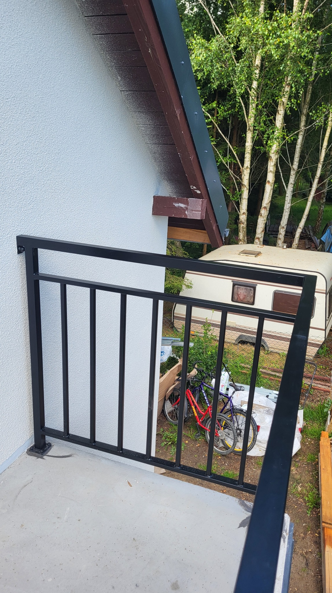 Czarna, metalowa balustrada balkonowa z pionowym wypełnieniem, zamontowana przy białej ścianie budynku. W tle widoczny przyczepa kempingowa i drzewa.