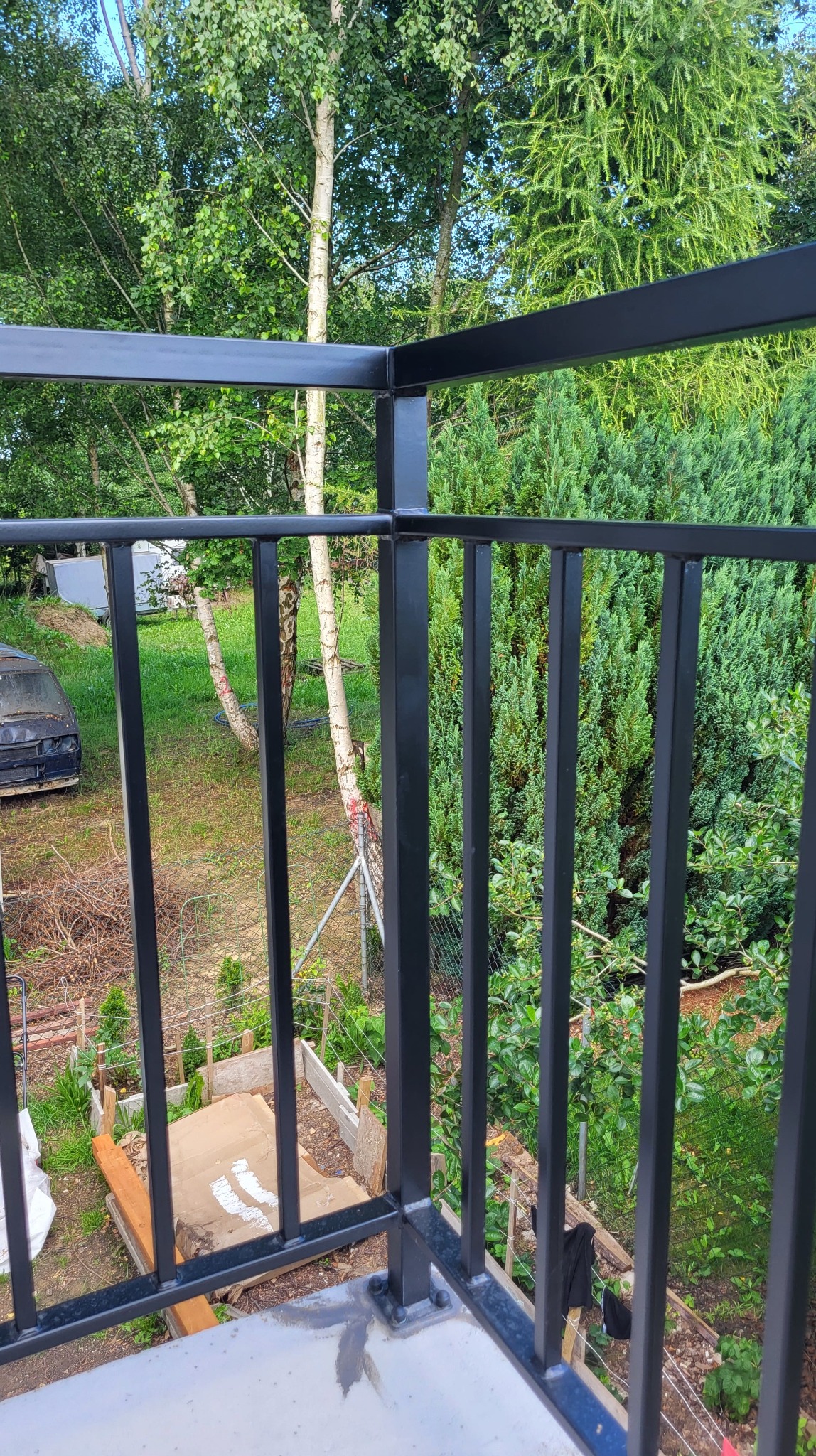 Czarna, metalowa balustrada balkonowa z pionowym wypełnieniem, widok z balkonu na zieleń, Koszalin. Elementy spawane, malowane proszkowo.