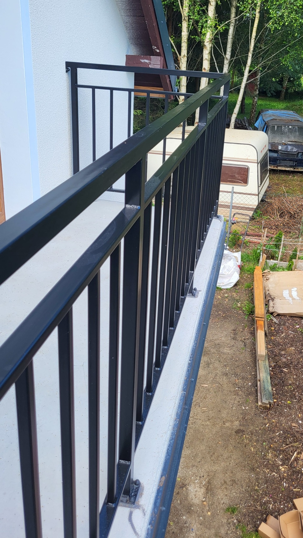 Czarna, metalowa balustrada balkonowa z pionowym wypełnieniem z profili, pomalowana proszkowo na mat. W tle widoczny przyczepa kempingowa i drzewa.