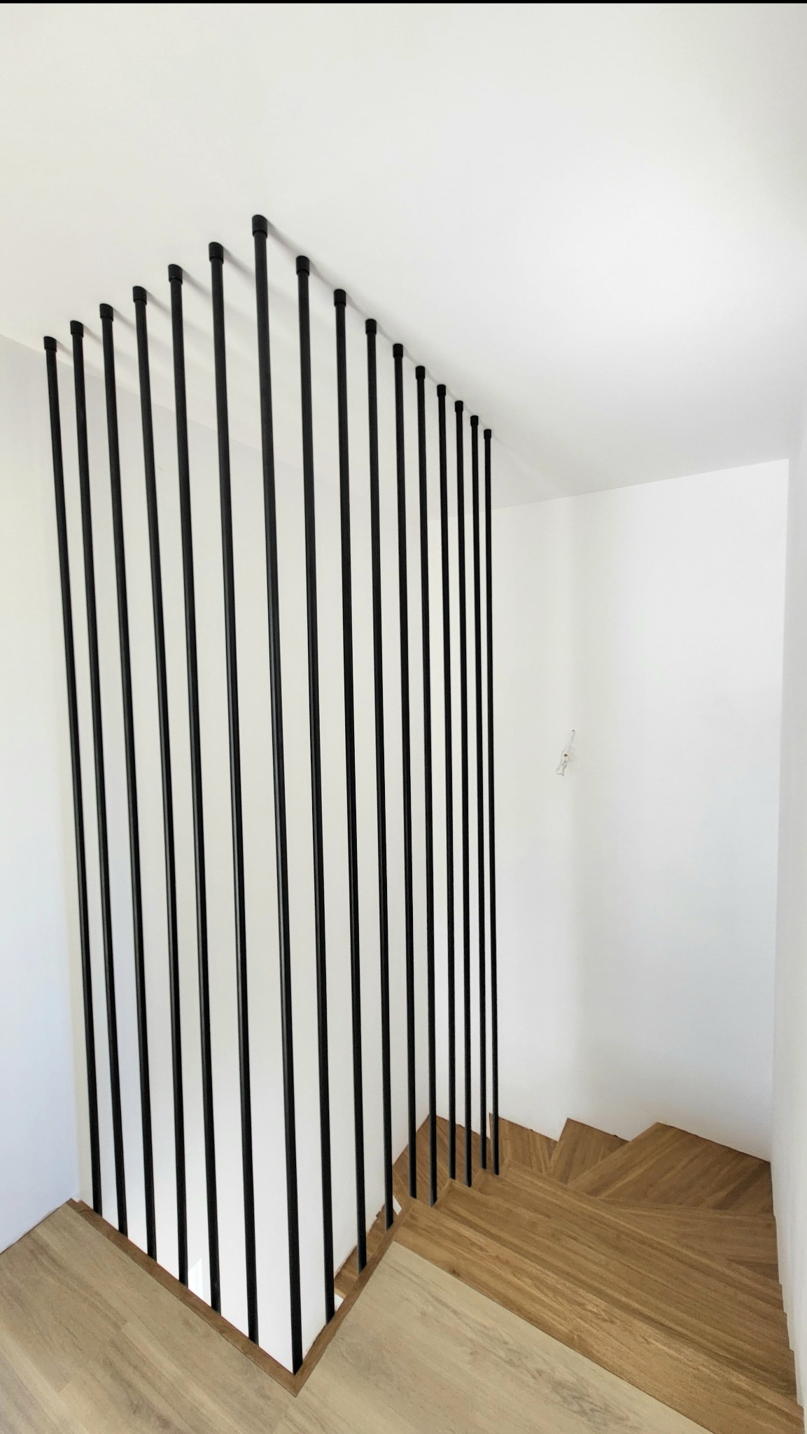 Nowoczesna balustrada schodowa z czarnych, pionowych rurek, mocowana do drewnianych stopni i sufitu, tworząca minimalistyczny design. Kontrast materiałów i kolorów.