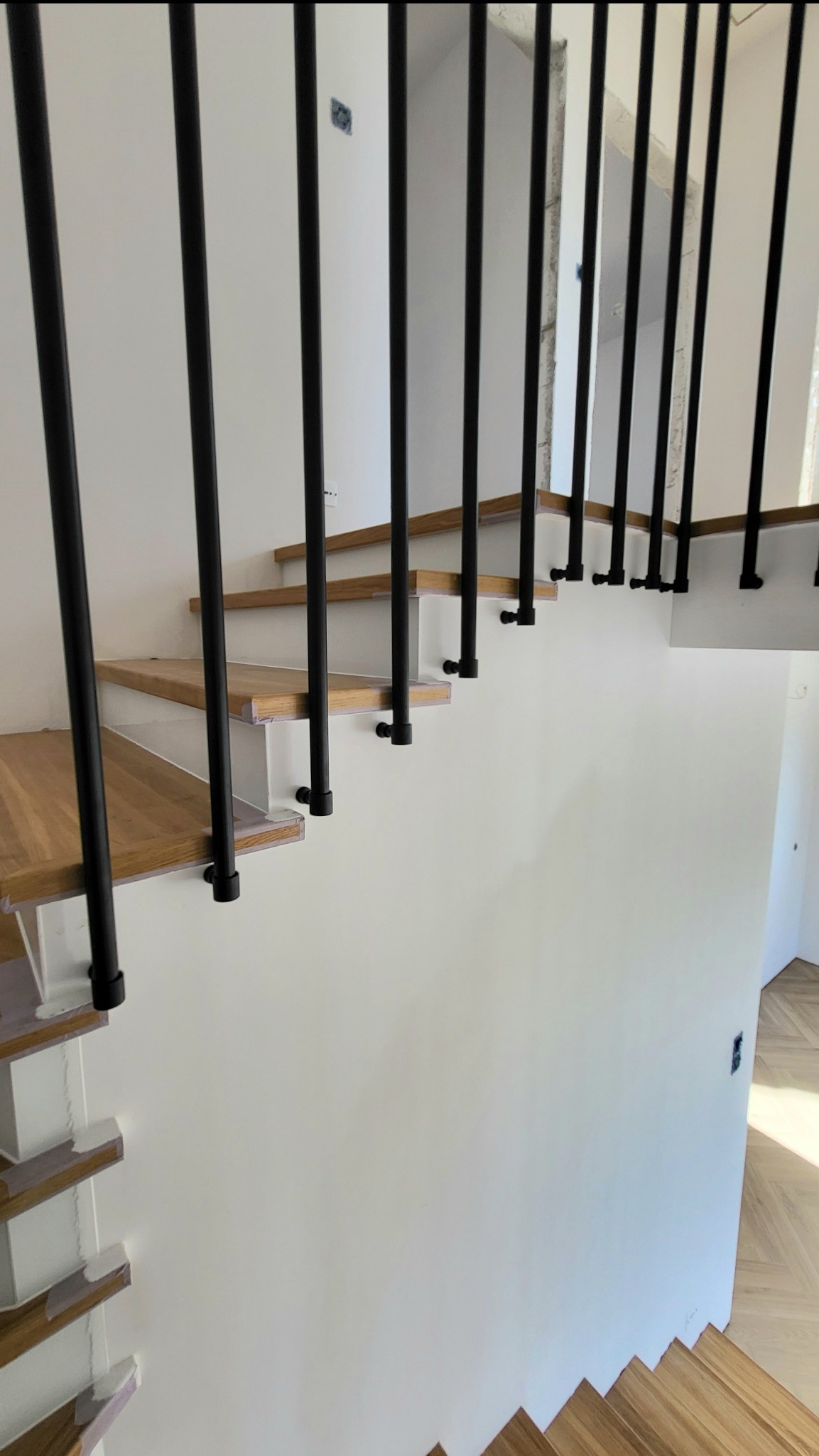 Schody drewniane z nowoczesną balustradą z czarnych, pionowych rurek. Minimalistyczny design, drewniane stopnie i białe ściany. Widok z dołu na balustradę.