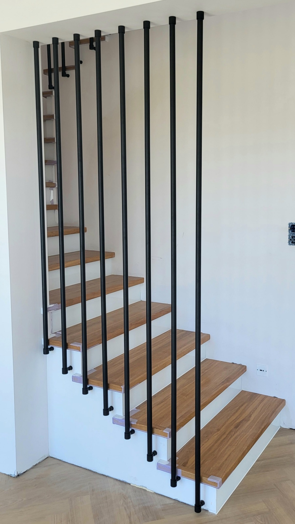 Nowoczesne schody z drewnianymi stopniami i czarnymi, metalowymi pionowymi prętami jako balustrada, mocowanymi do sufitu i boku schodów. Minimalistyczny design.