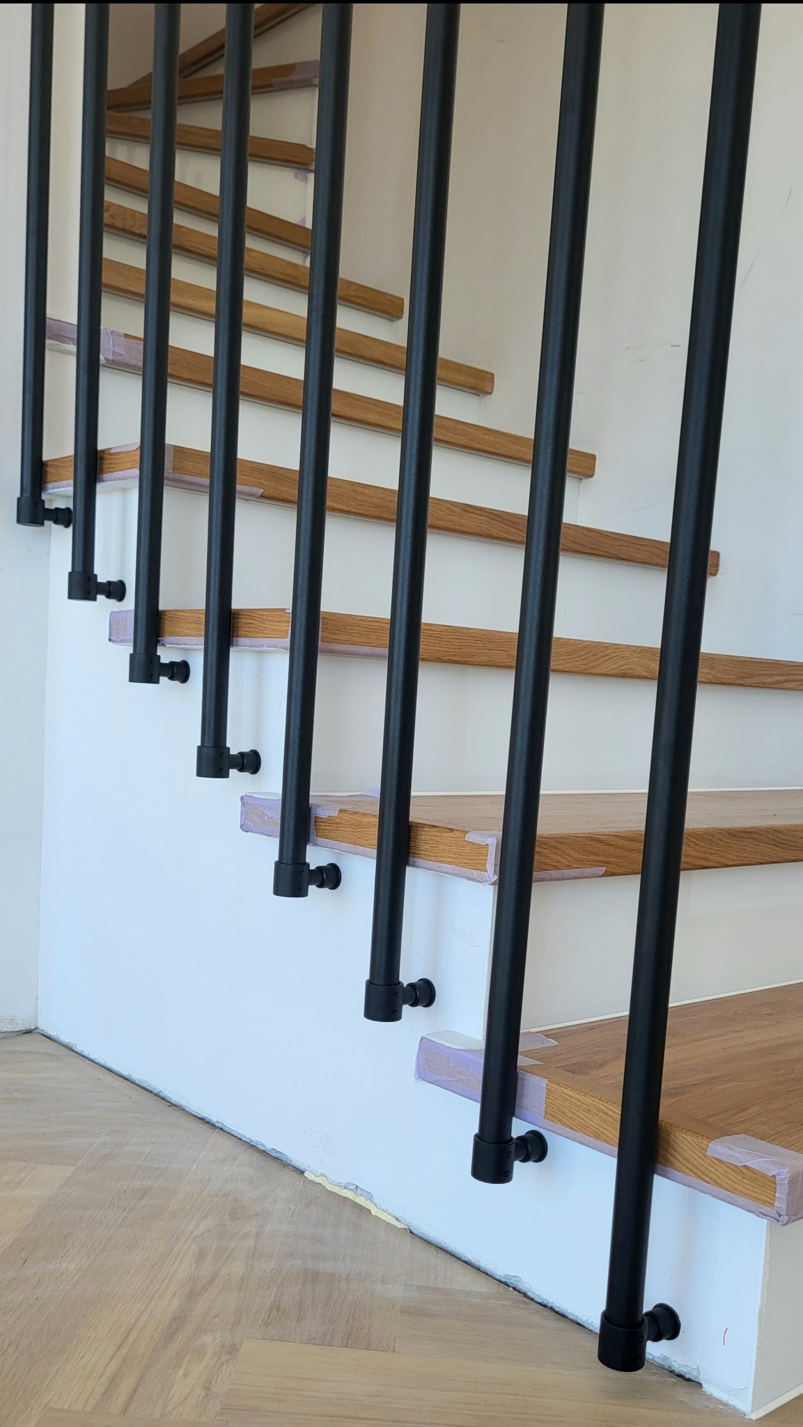 Nowoczesne schody z drewnianymi stopniami i czarną, metalową balustradą z rurek, mocowaną do boku schodów i sufitu. Kontrast materiałów i kolorów.