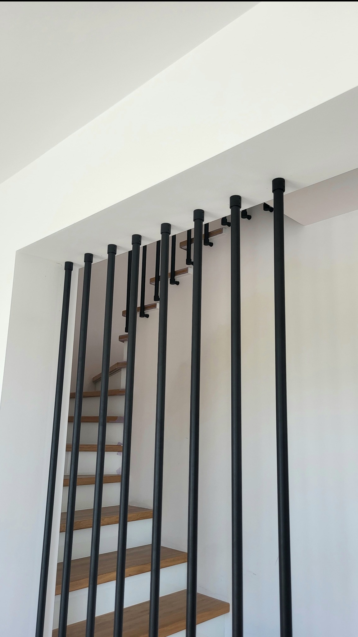 Nowoczesne schody z drewnianymi stopniami i balustradą z pionowych, czarnych rurek stalowych, mocowanych do sufitu i boku schodów. Minimalistyczny design.