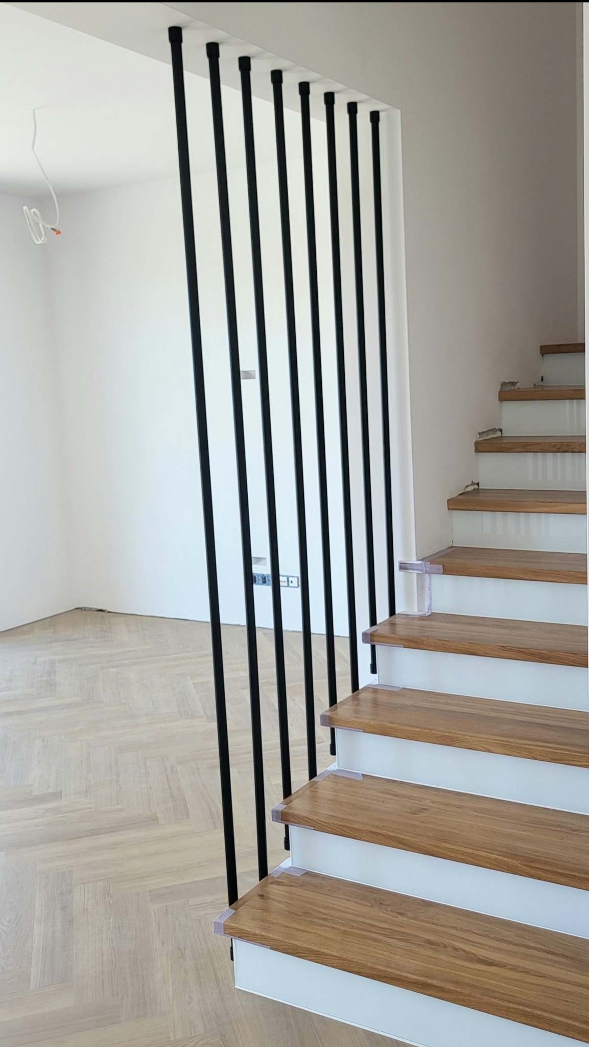 Nowoczesne schody z drewnianymi stopniami i czarną, minimalistyczną balustradą z pionowych rurek, mocowanych do stopnia i sufitu, na tle jasnego wnętrza.