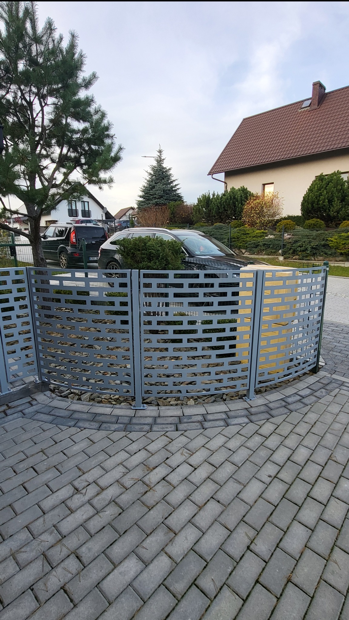 Szare, łukowe ogrodzenie z ażurowym wzorem, otaczające rabatę z krzewami. W tle dom z brązowym dachem i zaparkowane samochody. Kostka brukowa w szarym odcieniu.
