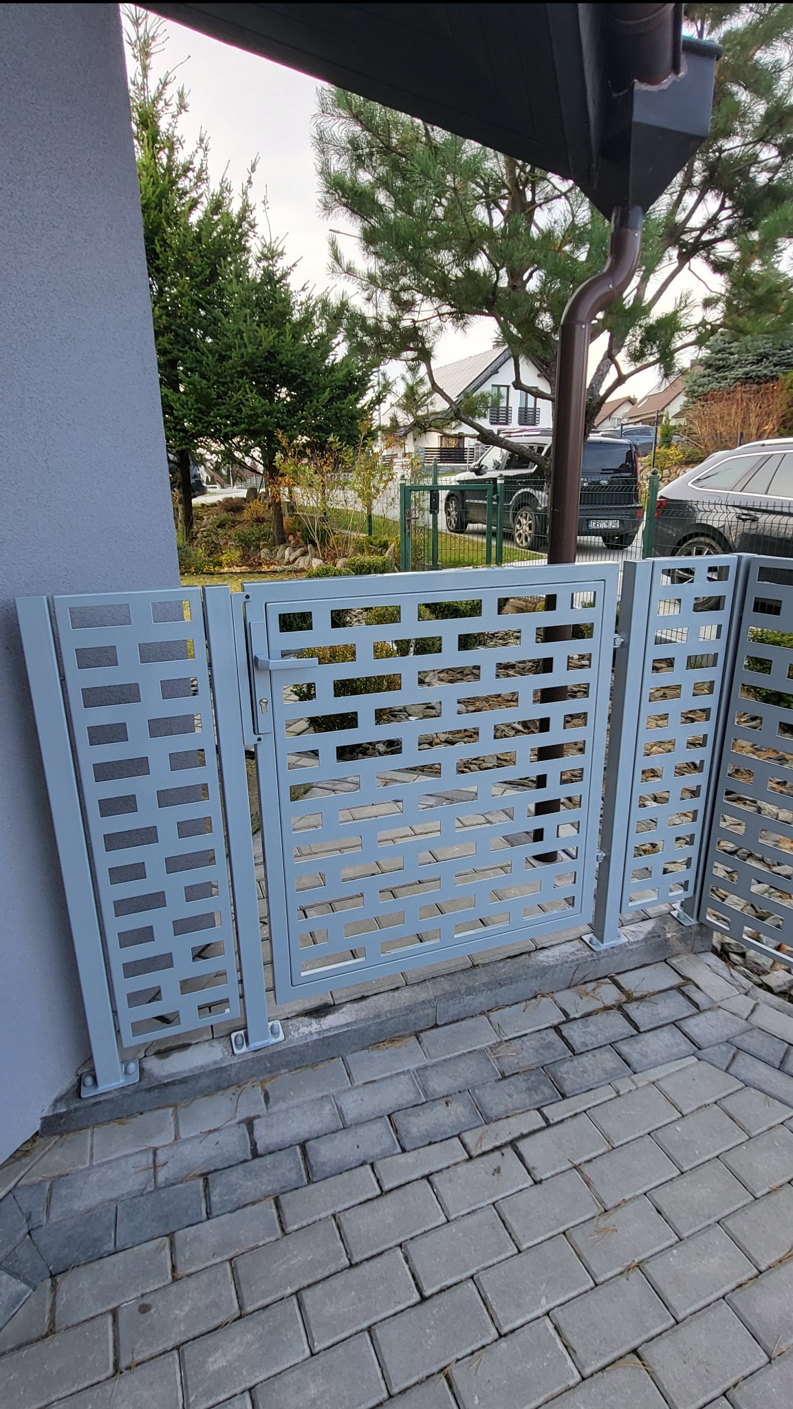 Szara, nowoczesna brama wjazdowa z ażurowym wypełnieniem z prostokątnych otworów, zamontowana na posesji z kostką brukową. Widok z bliska, w tle zieleń i budynki.