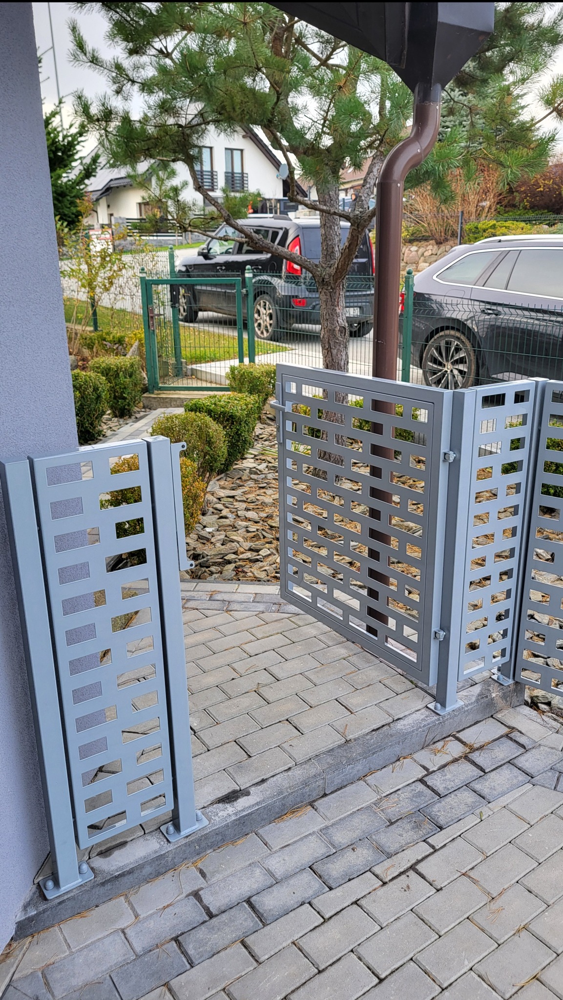 Nowoczesna, szara brama wjazdowa z ażurowym wzorem, zamontowana na posesji z kostką brukową i zielenią. Widoczne detale mocowania i wykończenia.