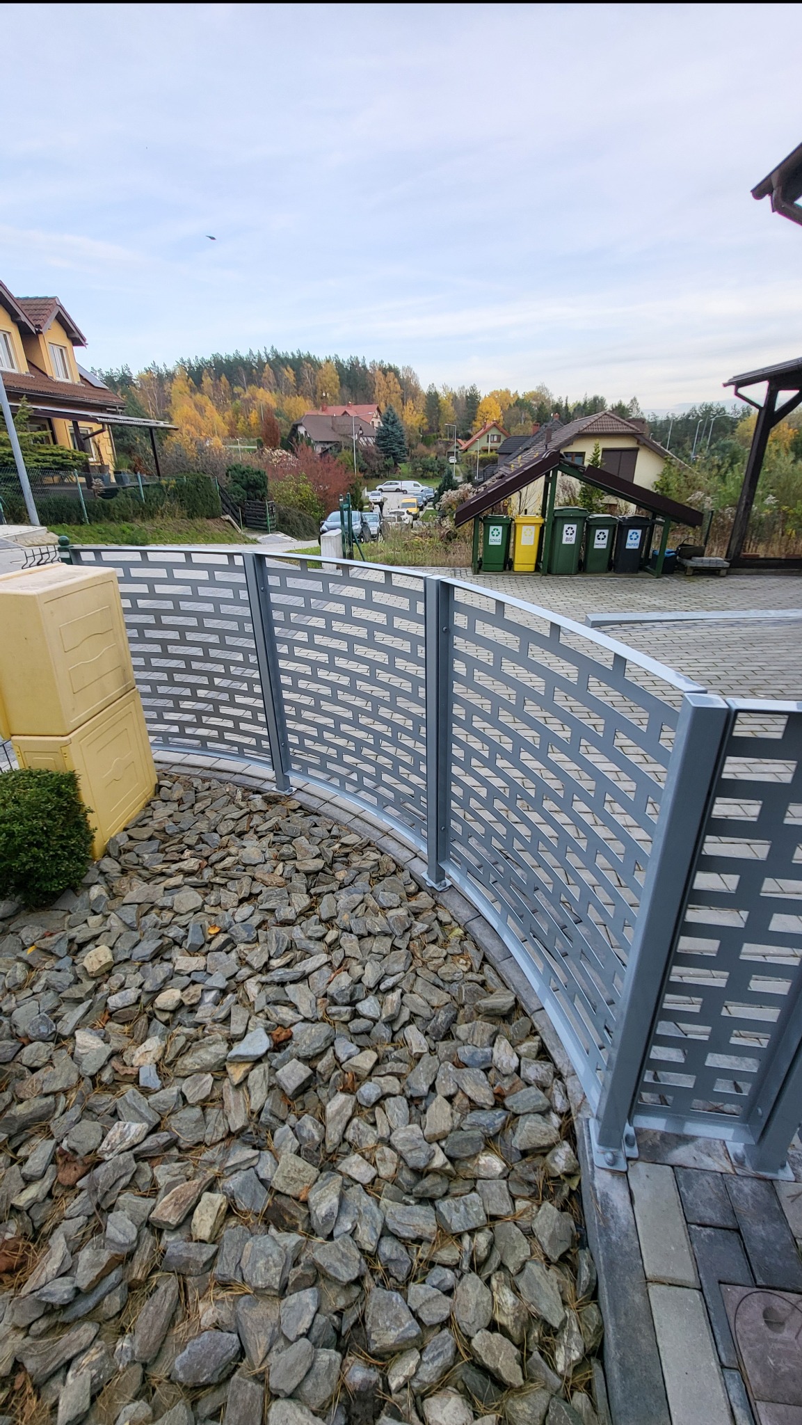 Łukowe, szare ogrodzenie z ażurowym wzorem, otaczające kamienny klomb. W tle domy i drzewa w jesiennych barwach. Widok z poziomu gruntu.