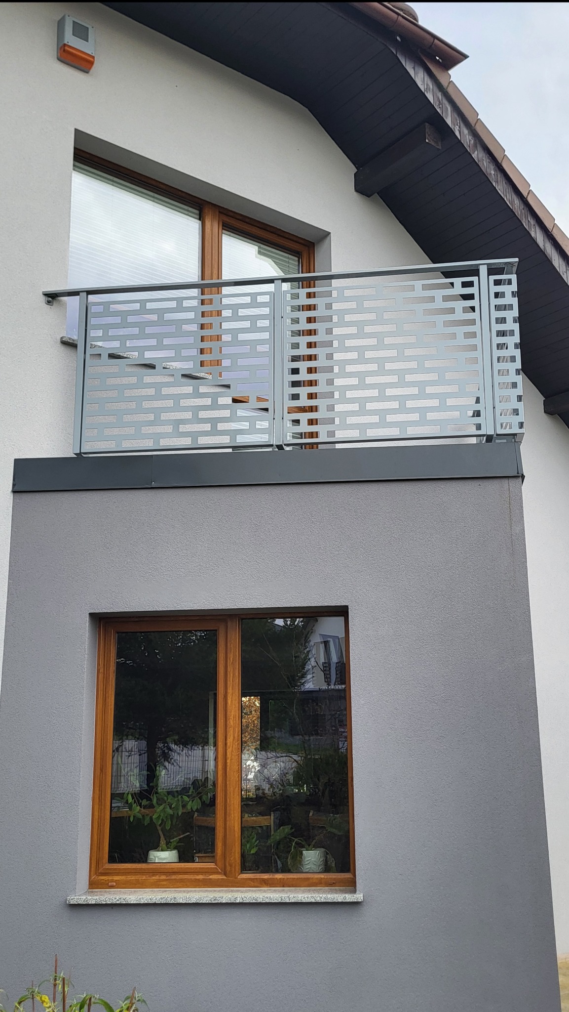 Szara, nowoczesna balustrada balkonowa z ażurowym wzorem cegiełek, zamontowana na balkonie domu z drewnianymi oknami. Widok z dołu, lekko z prawej strony.