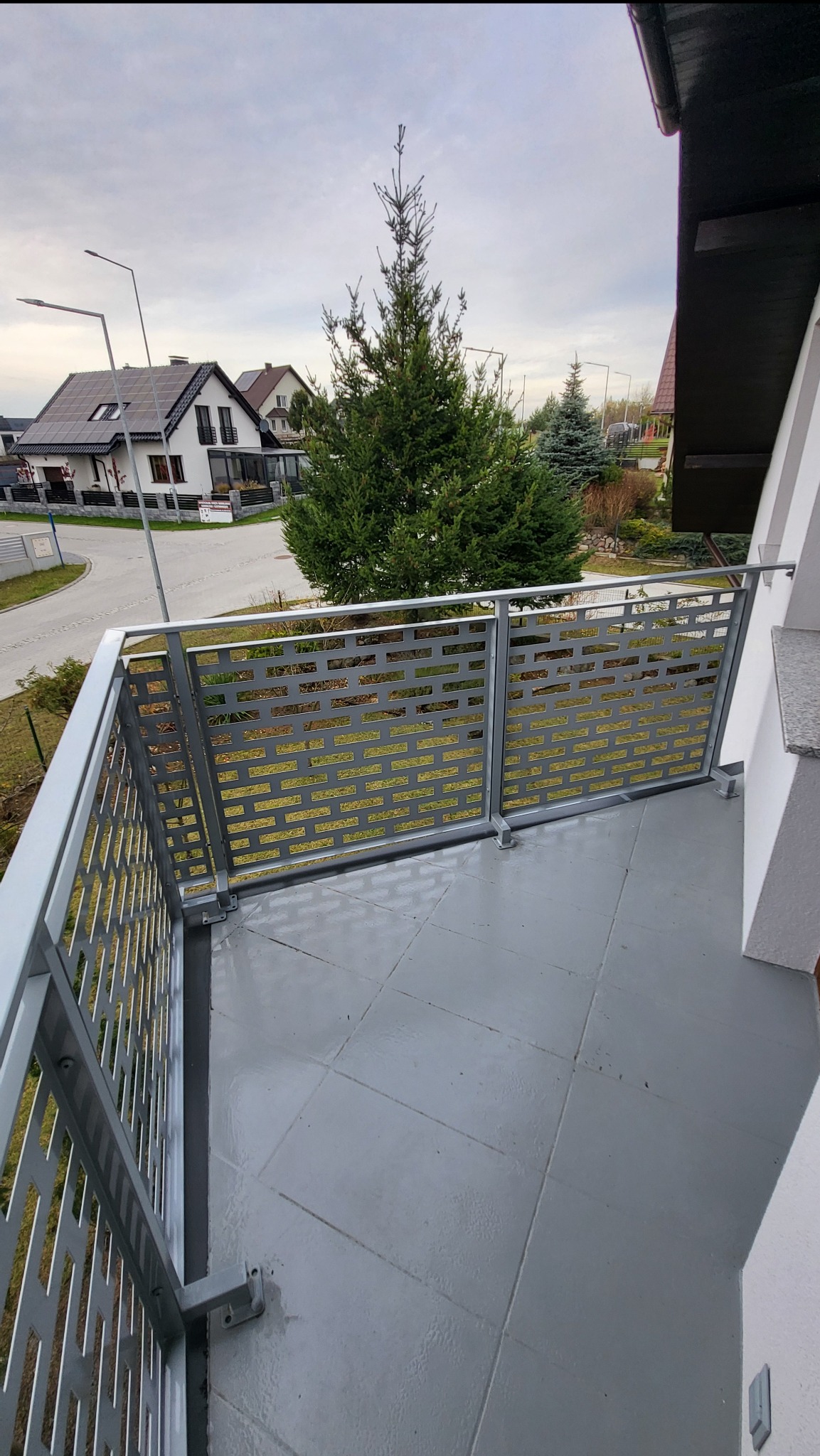 Szara, nowoczesna balustrada balkonowa z ażurowym wypełnieniem w kształcie cegiełek, widok z góry na tle zabudowy jednorodzinnej i zieleni.