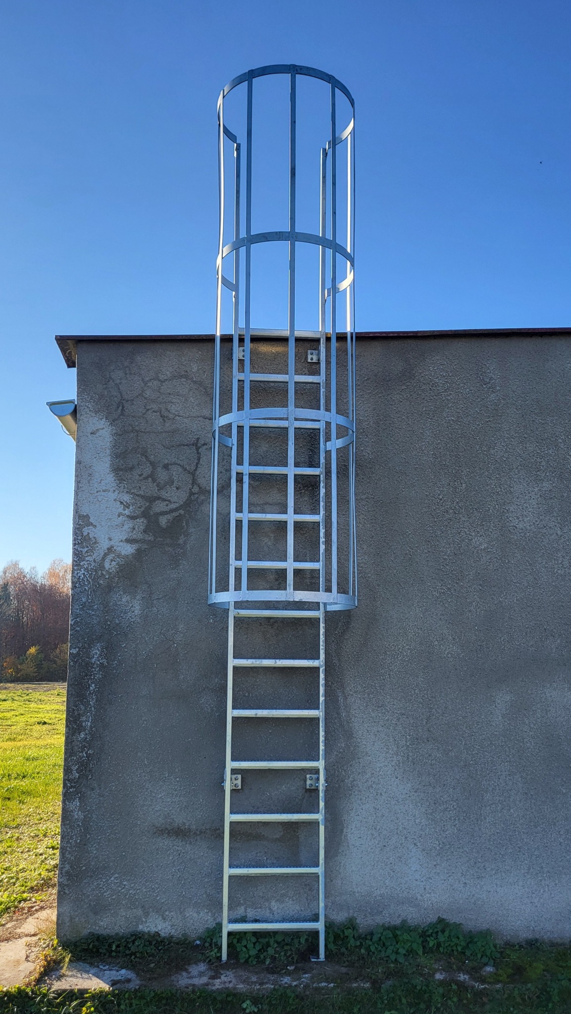 Metalowa drabina ewakuacyjna z koszem ochronnym, zamontowana na szarej ścianie budynku. Widok z dołu, błękitne niebo w tle. Drabina nowa, ocynkowana.