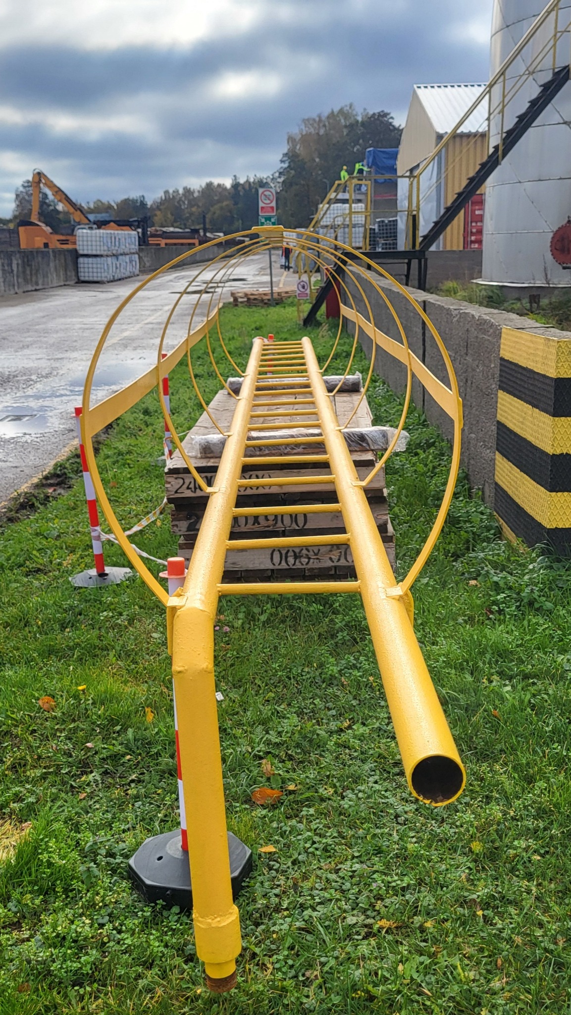 Żółta drabina z koszem ochronnym, leżąca na paletach na trawie, prawdopodobnie element instalacji ppoż. w tle budynki przemysłowe i dźwig.