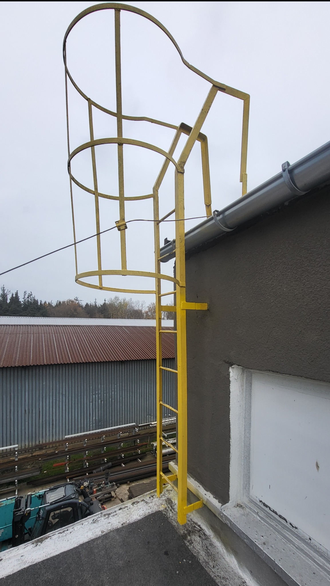 Żółta drabina ewakuacyjna z koszem ochronnym, zamontowana na ścianie budynku z szarą elewacją. Widok z dołu, na tle pochmurnego nieba i zabudowań przemysłowych.