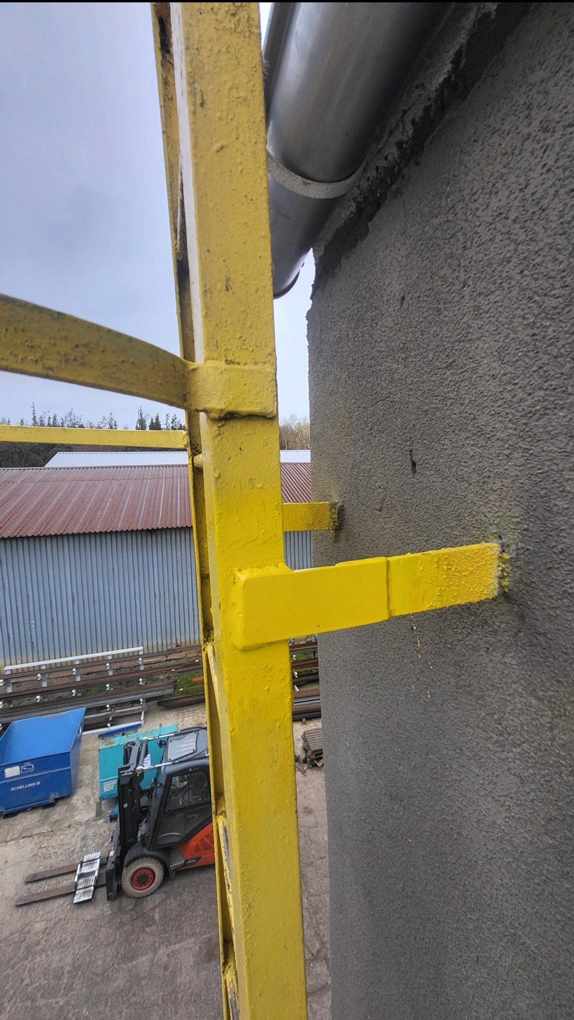 Żółta, metalowa drabina przymocowana do szarej ściany budynku. W tle widoczny wózek widłowy i magazyn z blachy falistej. Ujęcie z dołu, podkreślające wysokość konstrukcji.