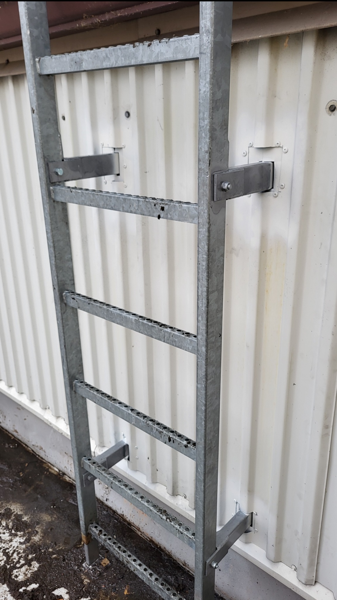 Metalowa drabina przymocowana do białej, falistej ściany. Widoczne mocowania i perforowane szczeble. Ujęcie z dołu, podkreślające pionowy charakter konstrukcji.