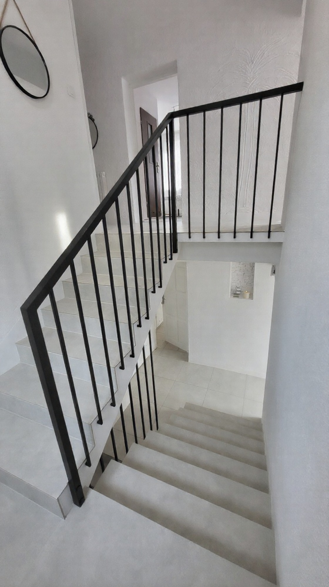 Minimalistyczne schody z czarną, metalową balustradą w jasnym wnętrzu. Tralki mocowane do ściany, dolna harfa z profilu. Nowoczesny design.