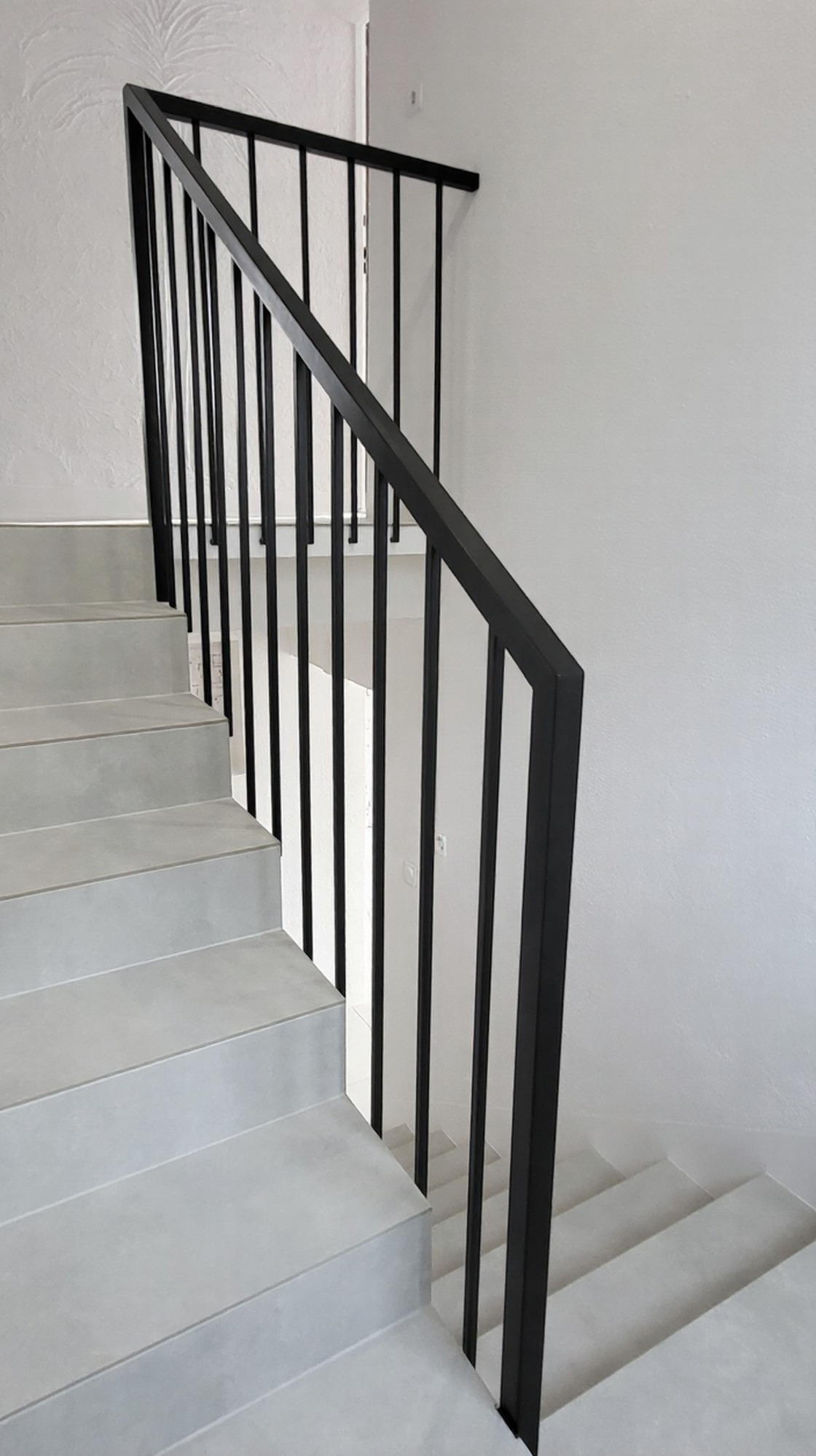 Minimalistyczna, czarna balustrada metalowa z pionowymi prętami na jasnych schodach. Nowoczesny design, proste linie, subtelne oświetlenie, białe ściany.